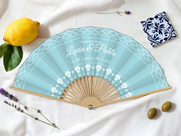 Abanico de boda personalizado «Orilla del mar», tela azul cielo e impresión en blanco, sobre una mesa de boda
