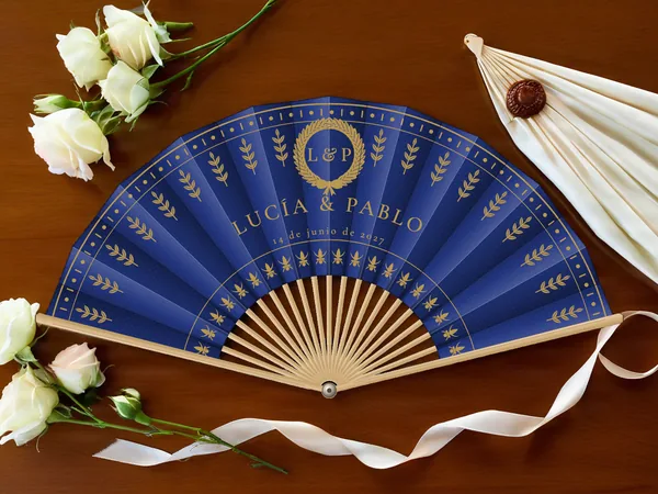 Abanico de boda personalizado «Laurel real», tela azul marino e impresión en oro, sobre una mesa de boda