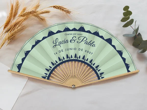 Abanico de boda personalizado «Abetos y montañas», tela verde agua e impresión en azul marino, sobre una mesa de boda